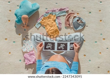 Pregnancy photo. Pregnant woman looking at first ultrasound photo of her baby with belly baby bump showing happily awaiting the birth of her child. Looking at baby clothing and toys on floor