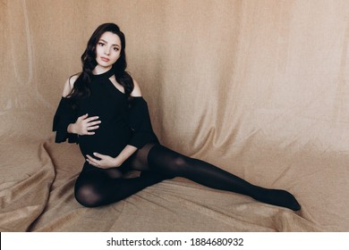 Pregnancy Photo Session In A Photo Studio With A Fabric Background In A Black Bodysuit And Tights