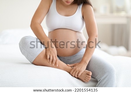 Pregnancy and problems with health concept. Unrecognizable pregnant woman in homewear sitting on bed at home, massaging her swollen foot, cropped shot, copy space