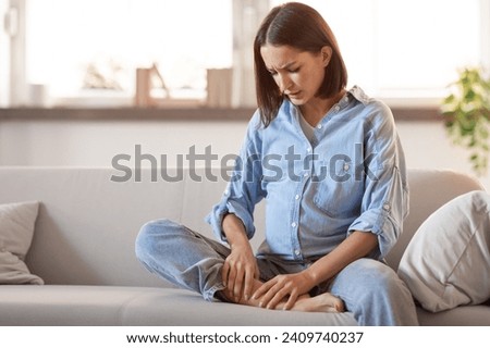 Pregnancy, problems with health. Young european pregnant lady in homewear massaging her swollen foot sitting on sofa at home indoor, having varicose vein issues during baby expectation