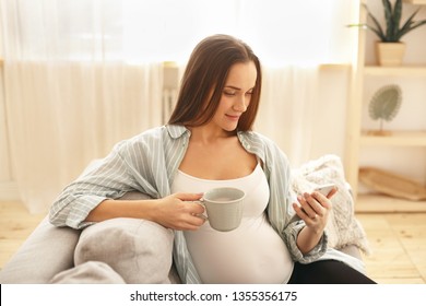 Pregnancy, Relaxation And Technology Concept. Beautiful Young Expectant Mother With Large Tummy Sitting Comfortably On Couch With Cup, Drinking Herbal Tea, Surfing Internet Using WiFi On Mobile Phone