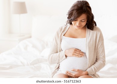 Pregnancy, Rest, People And Expectation Concept - Happy Pregnant Woman Sitting On Bed And Touching Her Belly At Home