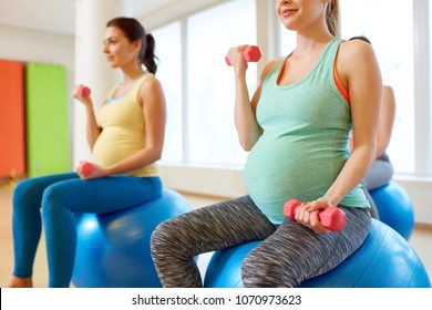 Pregnancy, Sport, Fitness, People And Healthy Lifestyle Concept - Group Of Happy Pregnant Women With Dumbbells On Exercise Ball In Gym