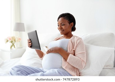 pregnancy, technology, people and expectation concept - happy pregnant african american woman with tablet pc computer in bed at home