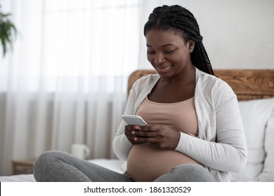 Pregnancy Tracker App. Happy Black Pregnant Woman Using Smartphone At Home, Smiling Expectant Mother Reading Information About Baby Development, Sitting On Bed With Cellphone, Relaxing In Bedroom