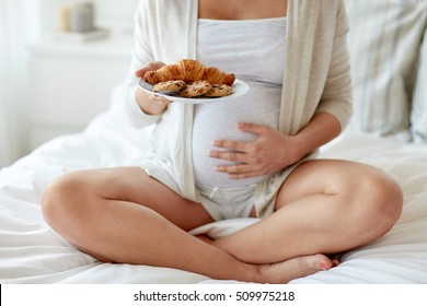 pregnancy, unhealthy eating, food and people concept - close up of pregnant woman with croissant buns and cookies sitting in bed at home