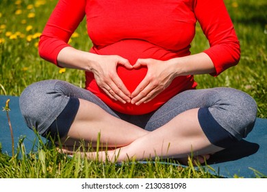 Pregnancy yoga exercise - pregnant woman doing asana Sukhasana easy yoga pose showing heart symbol with hands outdoors on grass lawn with dandelions in summer