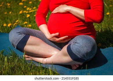 Pregnancy yoga exercise - pregnant woman doing asana Sukhasana easy yoga pose holding her abdomen outdoors on grass lawn with dandelions in summer