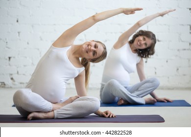 Pregnancy Yoga, Fitness concept. Two beautiful young pregnant yoga models working out indoor. Pregnant smiling fitness women sitting in yogic cross-legged pose at class. Prenatal sidebend in Easy Pose