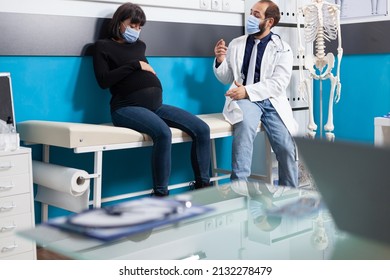 Pregnant Adult And Physician Doing Consultation During Covid 19 Pandemic. Patient Talking To Doctor With Face Mask About Pregnancy And Childbirth, Specialist Giving Medical Advice In Office.