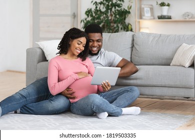 Pregnant african american couple having fun at home, using digital tablet together, expecting black lady showing her husband baby room interiors, sitting by couch at living room, empty space - Powered by Shutterstock