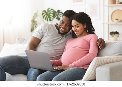 Pregnant african couple having fun at home, using laptop together, watching funny videos, empty space