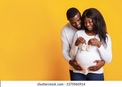 Pregnant african couple holding their future baby boots on wife belly and cuddling, yellow studio background, copy space