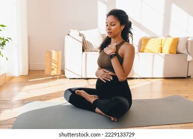 Pregnant african woman in lotus pose meditating on floor sitting on mat, doing pranayama breathing exercises for healthy pregnancy, preparing body for childbirth, putting hands of chest and belly - Powered by Shutterstock