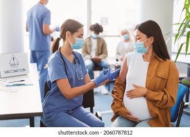 Pregnant Asian Lady Sitting At Doctor's Office Getting Modern Covid 19 Shot, Hugging Belly. Nurse In Medical Face Mask And Gloves Holding Syringe And Giving Female Patient Flu Vaccine Injection In Arm