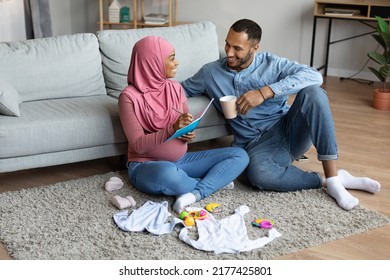 Pregnant Black Muslim Couple Making Checklist Of Necessities While Getting Ready For Childbirth, Happy Islamic Spouses Awaiting Baby, Relaxing On Floor In Living Room With Child Clothes Around