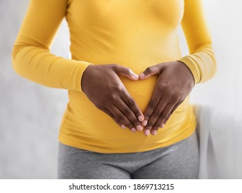 Pregnant black woman holding palms in heart shape on her big tummy, cropped. Unrecognizable african american expecting lady standing by window at home. Pregnancy and maternity concept