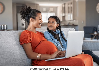 Pregnant Black Woman Working On Laptop While Sitting On Sofa And Smiling To Daughter. Expecting Mother And Girl Using Laptop. Happy Woman With Baby Bump And Daughter Working On Computer From Home.