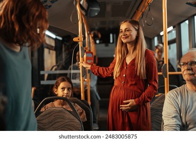 Pregnant blonde woman in a red dress happy as someone has given up their seat for her as she stands on a crowded public bus holding onto a handrail. - Powered by Shutterstock