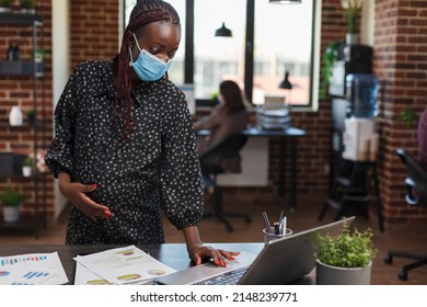 Pregnant businesswoman wearing virus protection mask in office using work laptop to review marketing strategy and proposed project ideas. Office worker reviewing accounting documentation. - Powered by Shutterstock