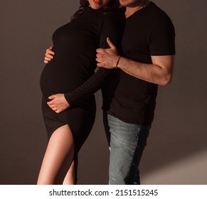 Pregnant Couple. Man In Black T-shirt And Jeans Hugs Pregnant Girl In Black Slim Dress On A Dark Studio Wall Background. Family Relationship. Pregnant Concept, Free Space