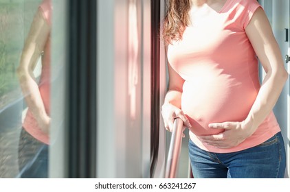Pregnant european woman is staying and holding her own tummy close to train window while she traveling by rail way and holding handrail inside the carriage at sunny summer day.