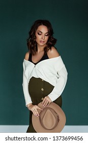 Pregnant european woman in white blouse and green skirt with beige hat in dark flat, stylish and happy pregnant woman in apartments, beautiful pregnant woman in dark studio