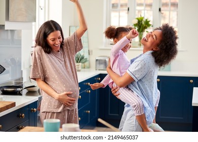 Pregnant Family With Two Mums Wearing Pyjamas Dancing In Kitchen With Daughter
