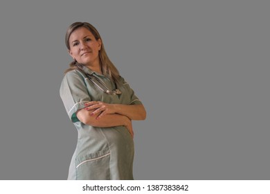 Pregnant female doctor or nurse stay in uniform with stethoscope and looks at camera smiling. Woman who is professional and future mother. Isolated on grey background