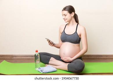 Pregnant female holding smartphone while sitting on exercise yoga mat at home at coronavirus time.