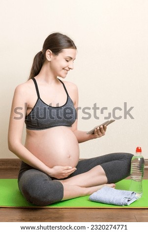 Pregnant female holding smartphone while sitting on exercise yoga mat at home at coronavirus time.