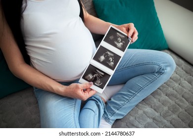 A Pregnant Female Shows Her Ultrasound Report And Her Unborn Baby. She Holds Ultrasound Baby Pictures With Both Her Hands In Front Of Her Big Pregnancy Stomach. A Woman Sits With Crossed Legs On Sofa.