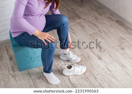 A pregnant girl with a big belly puts on sneakers on her feet. The concept of discomfort and inconvenience when bending over and putting on shoes. Convenient and comfortable shoes for pregnant women.