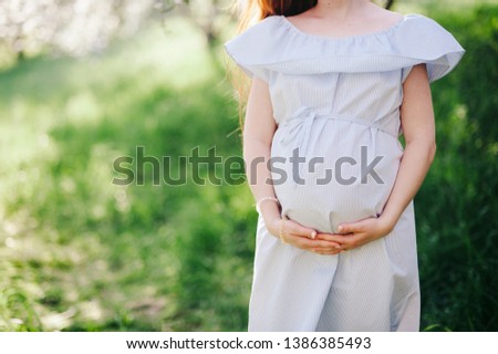 a pregnant girl in a blue dress is standing in a park with a green lawn and holding her belly