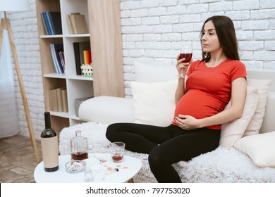 A pregnant girl drinks alcohol. She sits on the couch at home. She holds a glass of alcohol in her hand.