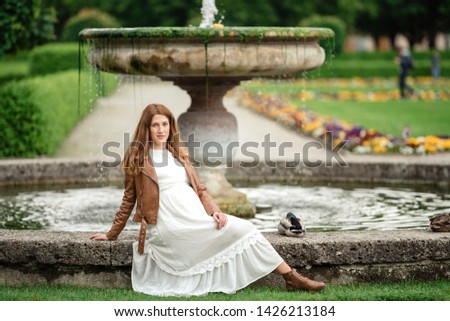 Pregnant girl near the fountain in full growth. A girl wearing a dress and a denim leather  jacket .