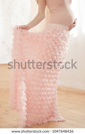 
A pregnant girl in a pink skirt is standing by the window, 