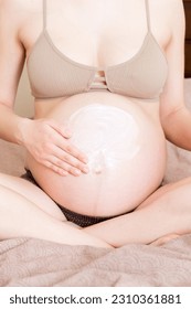 a pregnant girl sits at home on the bed and smears smile an anti-stretch mark cream on her stomach. Pregnancy, motherhood, preparation and expectation concept. - Shutterstock ID 2310361881