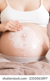 a pregnant girl sits at home on the bed and smears smile an anti-stretch mark cream on her stomach. Pregnancy, motherhood, preparation and expectation concept. - Shutterstock ID 2310361897
