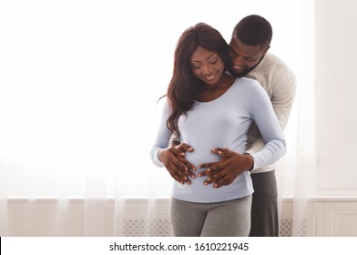 Pregnant happy black woman and her husband cuddling next to window at home, hugging belly, free space