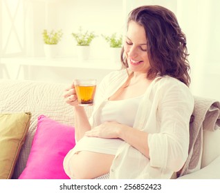 Pregnant Happy Smiling Woman Sitting On A Sofa And Drinking Herbal Tea. Mom Expecting Baby. Pregnant Woman Belly. Pregnancy. Beautiful Pregnant Woman. Maternity Concept.