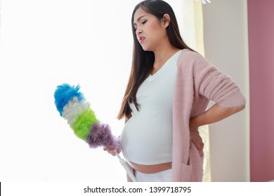 Pregnant Housewife Holding Feather Duster Suffering From Back Pain. Asian Pregnancy Doing Housework At Home.