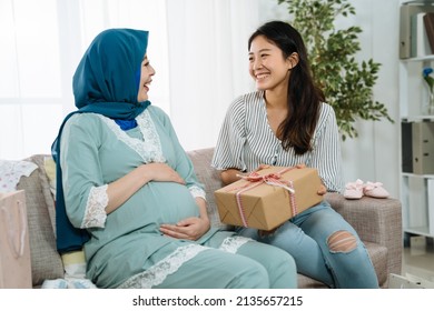 Pregnant Islam Woman And Asian Chinese Friend At Baby Shower Party Indoors In Home Living Room. Smiling Girl Sending Gift Expecting Unborn Baby In Big Tummy. Joyful Maternity Lady In Pajamas Laughing