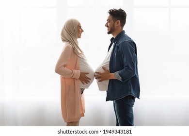 Pregnant islamic lady in hijab comparing belly sizes with her husband, having fun together at home. Joyful middle eastern guy with big fake tummy standing near window in front of her expectant wife