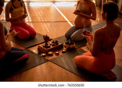 Pregnant ladies are sitting in circle and holding small cups. Little candles are in center. They meditating. Focus on tea ceremony