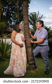 Pregnant Latin Couple Looking At Each Other With Tree In The Middle, In The Middle Of Tropical Forest With Lake