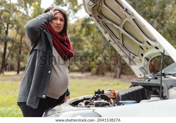 pregnant latina holds her\
head in front of her car\'s damaged engine and looks at the camera\
worriedly.