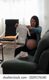 Pregnant Latina Woman Holding Up And Looking At Her Future Baby's Clothes.