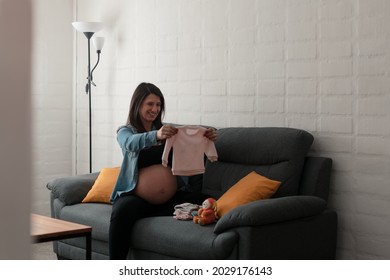 Pregnant Latina Woman Holding Up And Looking At Her Future Baby's Clothes.