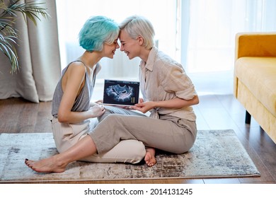 Pregnant lesbian couple with sonogram on floor in living room, side view portrait of two short-haired ladies in casual wear feeling love, happy to be future parents moms. copy space. lgbt, pregnancy